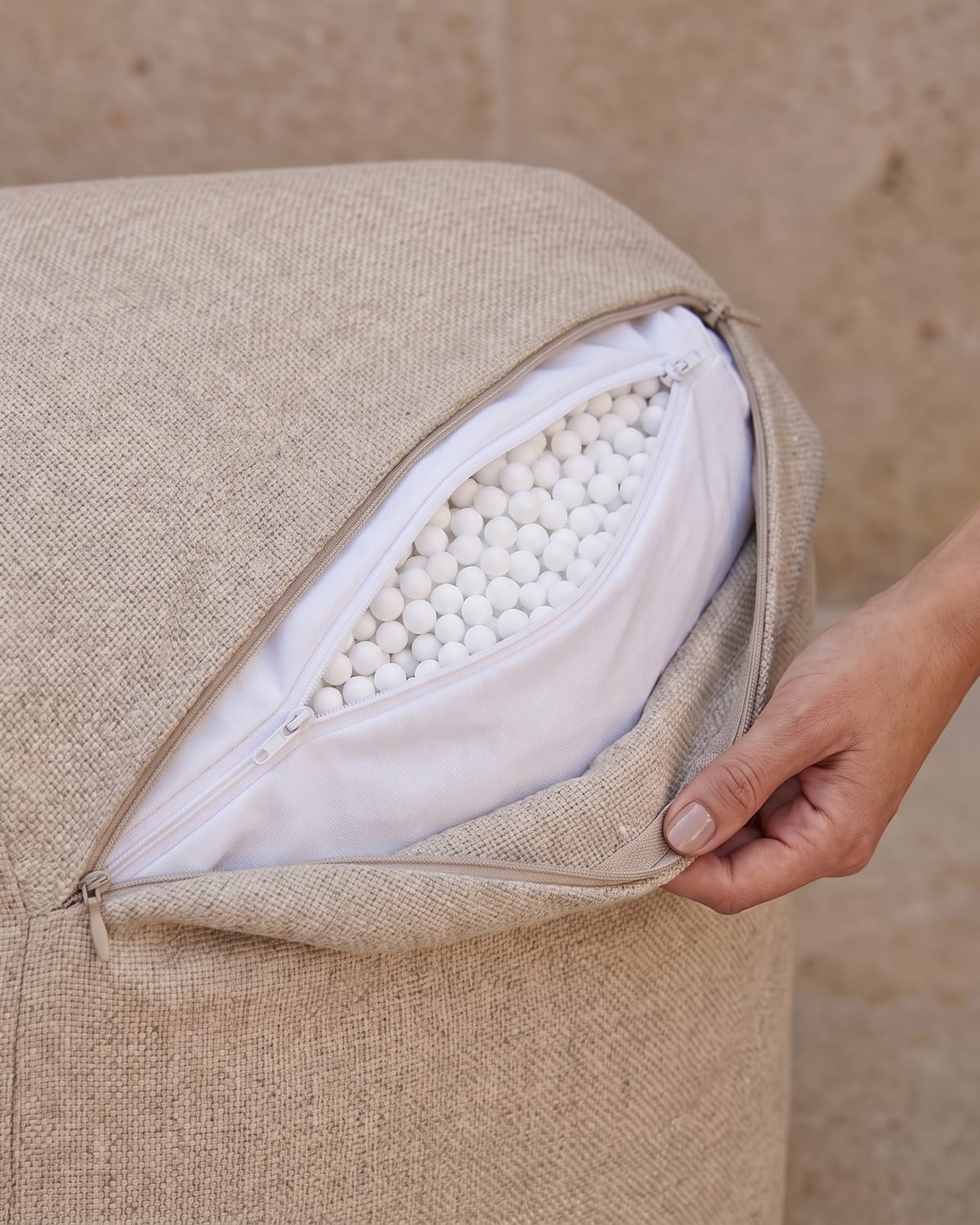 Hand opent de rits van een Nomad-fauteuil in beige outdoorstof. Daaronder zit een witte binnenzak met eigen rits, gevuld met ronde EPS-parels.
