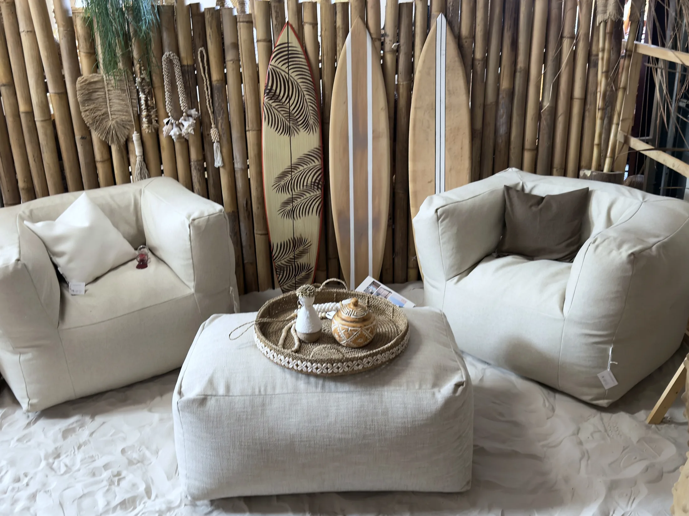 Twee linnen loungefauteuils met een poef en een rieten dienblad op de zandvloer bij CoCo homeliving, met surfplanken tegen een bamboewand